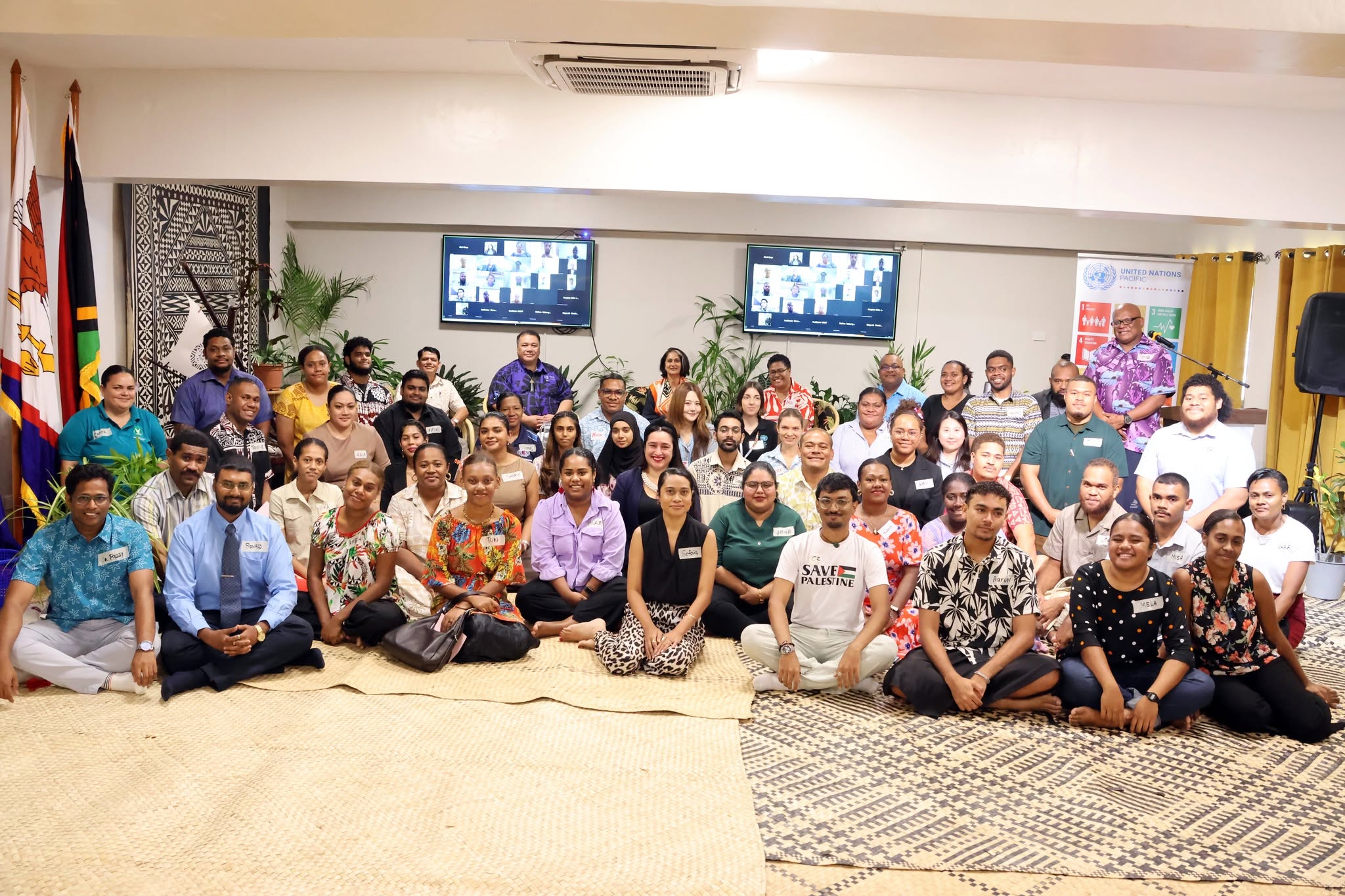 A large group photo of over forty Pacific youth leaders, civil society advocates, and UN representatives gathered in a brightly lit room for the Pre-COP31 Pacific Youth Talanoa. Participants are smiling, with several rows seated cross-legged on traditional woven mats on the floor and others standing in the background. In the back, two large television screens display virtual participants joining online, and a United Nations Pacific banner stands on the right.